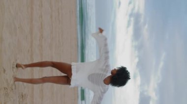 Mutlu kız beyaz bikinili sahili dikey seviyor. Gülümseyen Afro-Amerikan dansı tropikal kum kıyısında tek başına dinleniyor. Kaygısız siyah saçlı kadın okyanusta yalınayak yürüyor dikey olarak kendini özgür hissediyor.