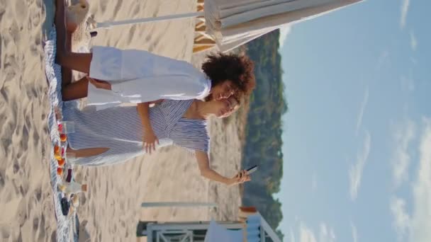 放宽心的女孩在海洋垂直处自拍 快乐的女同性恋夫妇在海滩休息 在社交媒体上发表文章 两个多民族的好朋友享受假期垂直的观点 友谊亲密性概念 — 图库视频影像