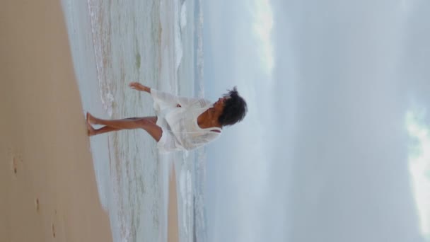 在海洋海岸线上行走的性感女人 快乐的无忧无虑的女孩穿着白色的衣服触摸沙滩捕捉海浪 松懈的梦幻般的非洲裔美国人垂直地享受着这个岛国的周末 和平度假胜地 — 图库视频影像