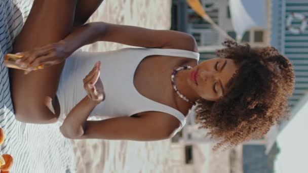 ビューティフルな女の子は 垂直ビデオを適用しています 海岸で日光浴をするスタイリッシュな女性 休暇中に紫外線保護クリームを施した垂直にアフリカ系アメリカ人マッサージボディ 休日のビーチリラックス — ストック動画