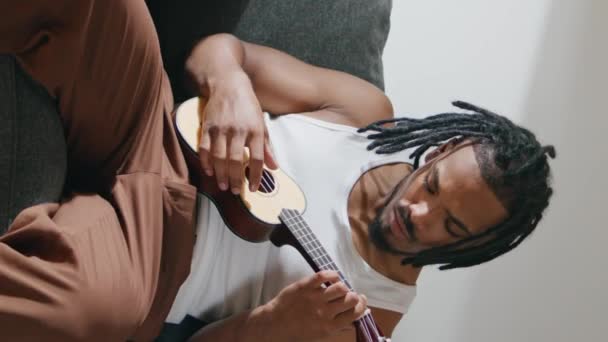 当代男人在轻便公寓垂直特写镜头下弹奏四弦琴 非洲裔美国人在舒适的沙发上提高音乐水平 发展从事吉他手活动的专业人员 爱好愉快 — 图库视频影像