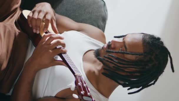有表现力的吉他手在国内垂直演奏四弦琴 非洲裔美国音乐家坐在沙发上触摸弦乐 黑头发的男人在公寓里放松地欣赏音乐 男人的手抽音 — 图库视频影像