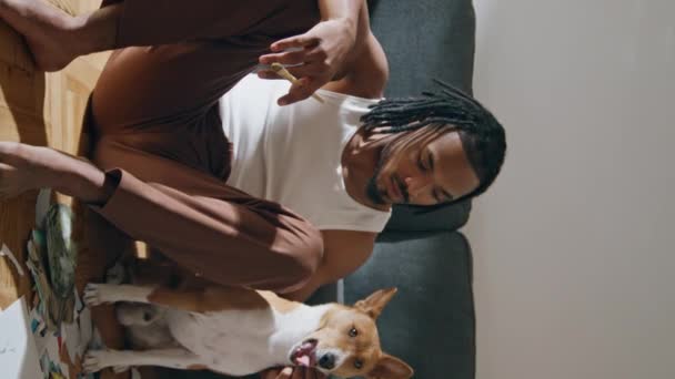 垂直にホームズームでペットをポーズするポジティブな男 アフリカ系アメリカ人男性が フロアの肖像画に座っているイヌを抱きしめている 笑顔のアーティストは 描いた後にリラックス 家畜と休む現代の画家 — ストック動画