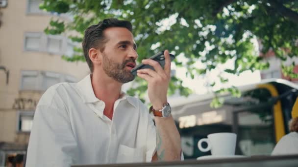 スタイリッシュなフリーランサーは 通りのカフェテリアに座って電話でボイスメッセージを指示します ラテン語の男性がダイナミックで音声通知を送信しています アウトドアでスマートフォンを使った自信のある大人のビジネスマン — ストック動画