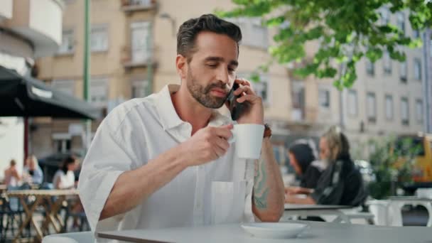 コーヒーハウスの携帯電話に電話する笑顔の従業員 ストリートカフェで会話をしながらエスプレッソを飲むポジティブなひげフリーランサー コーヒーを飲みながらコーヒーを飲むラテン系ビジネスマン — ストック動画