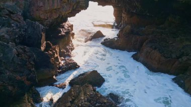 İnsansız hava aracı engebeli kayalıklarda köpüren dalgalı okyanus suyunu yavaş çekimde görüyor. Fırtınalı deniz dalgaları kayalık deniz kıyılarını yıkıyor güneşli bir yaz günü. Güçlü su tehlikeli volkanik kayalarda fokurduyor..