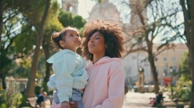 Güneşli sokakta durup park güzelini izleyen anne kızı. Mutlu hafta sonu yürüyüşlerinde gülümseyen Afro-Amerikan kadın güzel bir kızı elinde tutuyor. Tatlı çocuk güzel yeşil ağaçlara bakıyor..