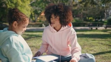 Anne kız doğada oturup hafta sonunun tadını çıkarıyor. Yakın çekim. Mutlu Afro-Amerikalı kadın aile pikniğinde Green Park 'ta oturan küçük kızla konuşuyor. Genç kıvırcık anne elinde kağıt kalem tutuyor..