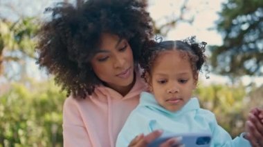 Afrika kökenli Amerikalı çocuk telefonda çizgi film seyrediyor. Neşeli genç anne, küçük kızına doğada eğlenceli uygulamalar gösteriyor. Kaygısız mutlu aile güneşli hafta sonunun tadını çıkar..