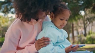 Küçük kız cep telefonunu izliyor yeşil doğada oturuyor ve kıvırcık annesi yakın çekim yapıyor. İyi dertsiz Afrika kökenli Amerikalı çocuk açık havada anne akıllı telefonunda çizgi film seyrediyor. Aile pikniğinde teknoloji.