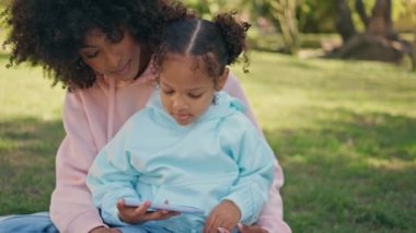 Çocuk, annesiyle birlikte doğa pikniğinde eğlenceli videolar izliyor. Rahat Afro-Amerikan annesi çimenlikte oturmuş akıllı telefon tutan küçük kızını seyrediyor. Aile hafta sonu telefonda parkta..