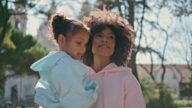 Kaygısız bir şekilde çocuğunu taşıyan gülümseyen bir kadın güneşli parka yaklaşıyor. Mutlu Afro-Amerikan annesi etrafı seyrediyor. Doğanın güzelliğinin tadını çıkarıyor. Kızını elinde tutuyor. Sevimli küçük kız annesiyle rahatlıyor..