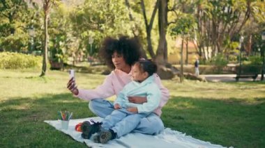 Afrika kökenli Amerikalı aile, akıllı telefon kullanarak doğada görüntülü görüşme yapıyor. Mutlu gülümseyen anne ve küçük tatlı kızı telefon kamerasına konuşuyor. Kaygısız kızlar yeşil çimlerde oturuyor güneşli çayırlarda