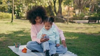 Anne kız telefonda oynuyor yeşil çimlerde güneşli çayırlarda oturuyor. Rahat bir Afrikalı Amerikalı kadın yaz doğasında küçük tatlı bir kıza akıllı telefon gösteriyor. Dışarıda cep telefonu kullanan mutlu bir aile.