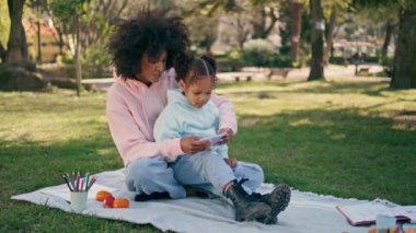 Anne kızı dışarıdaki akıllı telefon ekranına bakıyor. Mutlu Afrikalı Amerikalı kadın, Meadow 'da aile pikniğinde küçük kıza çizgi film gösteriyor. Gülümseyen anne, çocuğu telefonla eğlendiriyor.