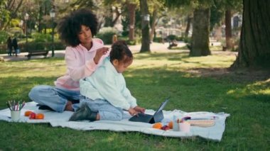 Anne, kızının piknikte otururken tablet oynamasını izliyor. Rahat Afro-Amerikalı bir kadın açık havaya odaklanmış şirin küçük bir kıza hayran. Güzel kıvırcık çocuk doğada klavye sekmesine basıyor..