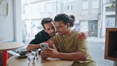 Romantik buluşmalarda akıllı telefon kullanan mutlu homoseksüeller. Yakın kafeteryada. Rahat eşcinsel çift cep telefonu uygulamasına bakıyorlar. Şirin çok kültürlü lgbt ailesi satın almaları telefonla seçiyor.