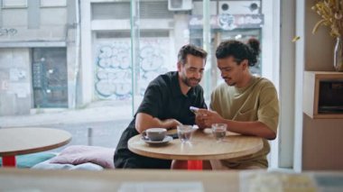 Küçük restoran masasında oturan akıllı telefon görünümlü eşcinsel çift. Genç Latin adam cep telefonunda sakallı ortağının fotoğrafını gösteriyor. Sosyal medyayı cep telefonundan izleyen mutlu çoklu etnik erkekler.