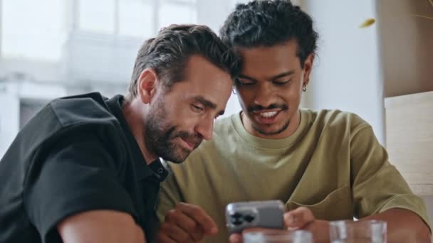 複数の同性カップルは カフェの近くに座っている携帯電話のスクリーンを見ながら笑います 2人の同性愛者が面白いビデオを見ている カフェテリアでスマートフォンを使用したハッピーケアフリー Lgbtq パートナー — ストック動画