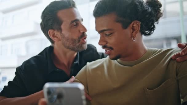 Lgbt合伙人坐在街头咖啡店附近讨论网上购物 笑多种族同性夫妇看智能手机屏幕 快乐的同性恋男人享受在一起的时光 — 图库视频影像