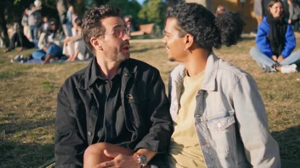 Lgbtファミリーは芝生公園に座り 笑顔で近づいていた 同性愛のゲイカップルを愛し 晴れた自然の中で夏の週末を過ごしています 混合レーストランスジェンダー関係性ジェンダーの許容コンセプト — ストック動画