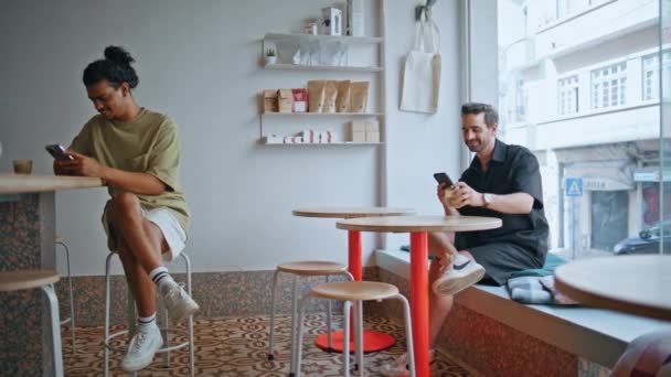 カフェテリアに座っている2人の男がスマートフォンの画面を見ています 快適なコーヒーハウスのインテリアで個別にリラックスしたリラックスしたマルチエスニックカフェ 携帯電話でアロマティックコーヒーを飲むカップル男性 — ストック動画