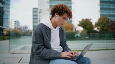 Sokak kapanışında çalışan serbest çalışan genç bir bilgisayar. Kıvırcık çocuk dışarıda laptopta sosyal medyada sohbet ediyor. Dijital aygıtta içerik oluşturan sakin blog yazarı. Modern binalar bölgesinde oturan hippi.