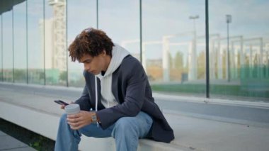 Dışarıda akıllı telefonlardan konuşan genç bir adam. Kıvırcık saçlı adam cep telefonuyla konuşuyor. Şehir merkezinde tek başına oturuyor. Cam bina arka planında kahve fincanı tutan rahat bir genç. Modern blogger mesajlaşma telefonu