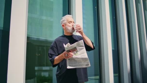 ビジネスマンがダウンタウンでコーヒーを飲む 思慮深いシニア読書新聞は市街地で朝のルーチンを楽しんでいる 現代のオフィスビルで休憩する灰色の髪の男性幹部 — ストック動画