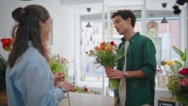 ブーケショップで笑顔の顧客購入の花の構成 休日のお祝いに花のアートアレンジを購入するカーリーヘア男性を閉じます 花の植物店のオーナーが花を売っています フローリスト販売コンセプト — ストック動画