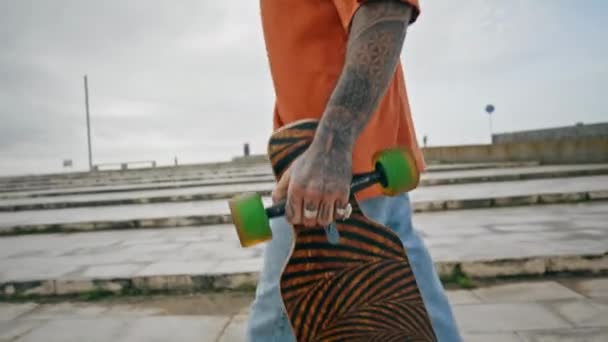 不知名的纹身男子滑板与现代长板在街上行走 年轻的长板手提着滑板在城市的背景下前行 难以辨认的滑板手在多云的户外漫步 — 图库视频影像