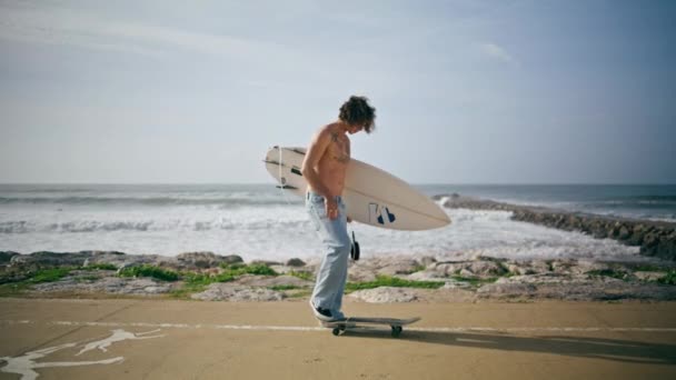 年轻男子冲浪运动员在美丽的夏季海洋特写附近的路上滑板 迷人的时髦男人拿着白色的现代冲浪板看相机带着微笑 卷曲纹身滑板手去冲浪 — 图库视频影像