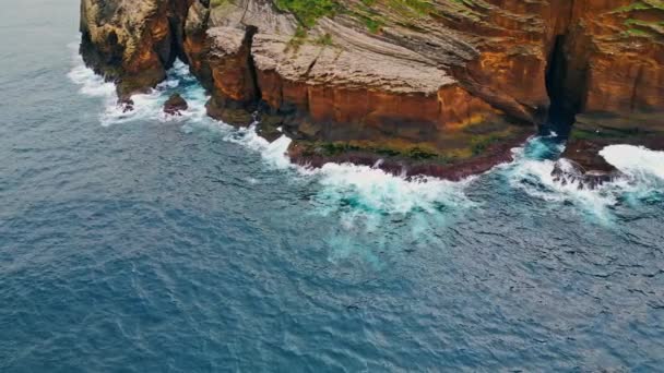岩の海岸線を衝突する発泡波 夏の日に野生の海がサンゴ礁の岩崖を洗っている 荒れた島の石に衝突する強力な海水のドローンショット 美しい海岸の手つかずの自然の概念 — ストック動画