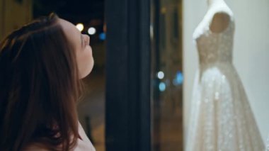 Gelinlik görünümlü gelinlik akşamları gelinlik mağazasının yanında duruyor. Genç hayalperest kadın vitrindeki beyaz pahalı elbiselere bakıyor. Mutlu kız gelinlik seçiyor..