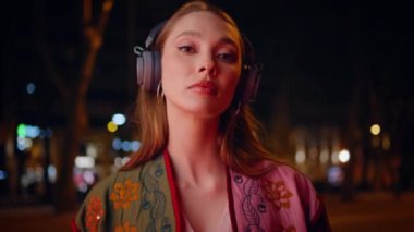 Kulaklık takan bayan hippi, gece kentinde kameraya özgüvenli bir şekilde bakıyor. Kablosuz kulaklıklarla müzik dinleyen muhteşem bir kadın akşam caddesinde duruyor. Moda kızı kulaklıkla poz veriyor.