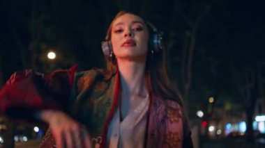Gece gezen kulaklıklı neşeli kız yaklaş. Gecenin geç saatlerinde kablosuz kulaklıkla müzik dinleyen mutlu ve rahat bir kadın. Tasasız moda bayan modern kulaklıkla sesin keyfini çıkar..