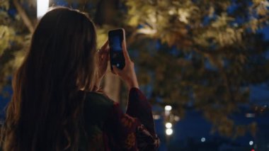 Akıllı telefondan gece parkının fotoğrafını çeken kız. Çekici genç bir kadın Alacakaranlık 'ta tek başına fotoğraf çekiyor. Rahat bayan cep telefonuyla karanlık şehri kaydediyor..