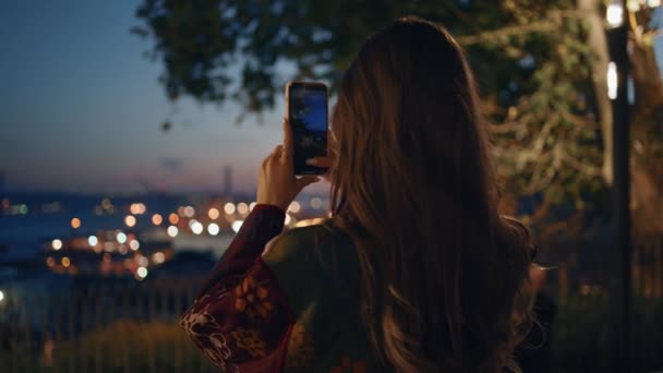 在智能手机上拍摄夜景的女人站在观景台旁边 年轻女性博主用手机拍摄全景城市夜灯的背景图 观光客夫人拍照 — 图库视频影像