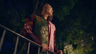 Gece parkında ayakta duran kadın korkuluklara yaslanıyor. Alacakaranlık vaktinde yeşil ağaçların altında poz veren gülümseyen rahat kız. Karanlık şehir bahçesinde kaygısız bayan bekliyor. Akşam hayatı konsepti
