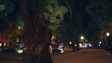 Kaygısız kız alacakaranlık parkında yürüyor ve kameraya gülümsüyor. Rahat maceraperest kadın karanlık şehirde akşam gezintisinin tadını çıkarıyor. Sokak lambaları altında poz veren modaya uygun bayan turist..