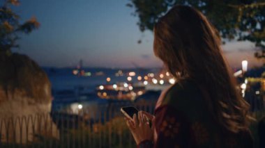 Park gözlem güvertesinden panoramik fotoğraf çeken bir kadın. Çekici genç kız turist akıllı telefondan video gece görüntüsü kaydediyor. Karanlık şehrin fotoğrafını çeken blogcu.