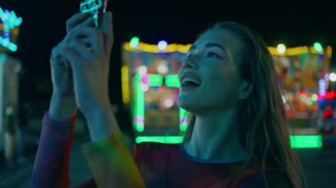 Akıllı telefonu elinde tutan heyecanlı kız video kaydını kapatıyor. Tatil gecesinde neşeli kadın dinlenme, lunaparkı. Gülümseyen mutlu genç neon renkli atlıkarıncada fotoğraf çekerken eğleniyor.