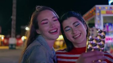 Neşeli en iyi arkadaşlar yaz akşamı lunaparkında selfie çekiyorlar. Gülümseyen modeller lunaparkta dinlenip cep telefonu fotoğrafları çekiyorlar. Muhteşem mutlu kız arkadaşlar birlikte eğleniyorlar. Kaynaşma anları boş zaman