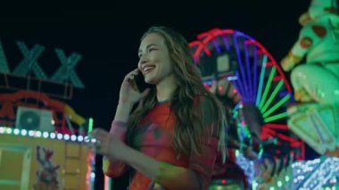 Lunaparktaki hücrede konuşan güzel bir kız. Gülümseyen kadın renkli parlayan dönme dolapta akıllı telefon yazmayı bitiriyor. Rahatlamış genç arkadaşları bekliyor, lunaparkta tatilin tadını çıkarıyorlar..