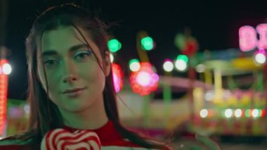Güzel bir lolipopla poz veren güzel bir kız. Gülümseyen genç lunaparkta yürüyüp akşam buluşmasının tadını çıkarıyor. Güzel bir kadın bulanık renkli ışıklara bakar. Hafta sonu eğlence konsepti