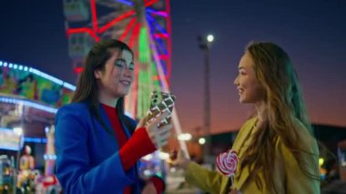 Neşeli kızlar lunaparkta selfie çekerler. Mutlu en iyi arkadaşlar akıllı telefondan fotoğraf çeker. Aydınlanmış lunaparkta akşamın tadını çıkarırlar. Göz kamaştırıcı kız kardeşler gece birlikte dinlenirken iyi eğlenceler..