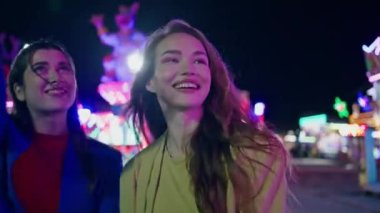 Neşeli kızlar gece eğlence parkında yürüyorlar. Mutlu en iyi arkadaşlar yaz festivali eğlencesinde birlikte gülerler. Muhteşem gülümseyen mankenler neon ışıklı atlıkarıncada tatilin tadını çıkarıyorlar.