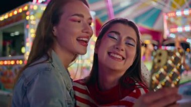 Komik arkadaşlar cep telefonlarının fotoğrafını çekiyorlar. Neşeli kızlar neon ışıklarda birlikte eğleniyorlar. Gülümseyen kız arkadaşlar lunaparkta hafta sonunun tadını çıkarıyor..