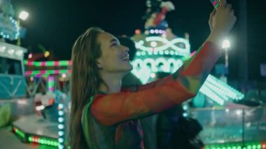 Lunaparkta selfie çeken mutlu kız. Gülümseyen genç, neon ışıklı lunaparkta cep telefonu kullanarak sosyal medyada eğleniyor. 