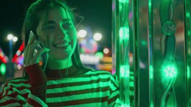 Neşeli kız neon ışıklarıyla hücreden konuşuyor. Mutlu genç el sallayan arkadaş lunapark eğlencesi. Güzel gülümseyen bir model cep telefonuyla akşamın tadını çıkar. Dostça bir hoş geldin jesti.